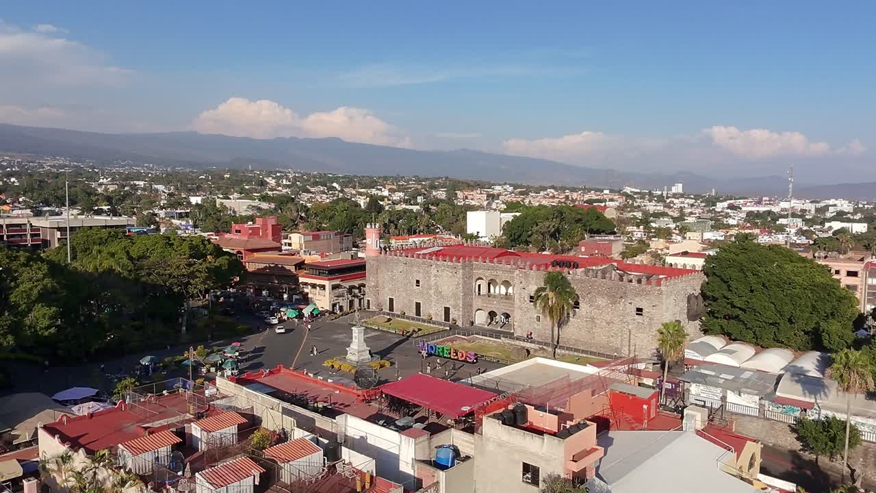 Backward drone aerial shot of Palacio de Cortés, colorful Morelos letters, central plaza with Carlos Pachecho statue, and sprawling Cuernavaca city with mountains in the background.