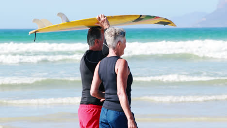 Una-Pareja-Mayor-Llevando-Una-Tabla-De-Surf-Sobre-La-Cabeza-Mientras-Caminaba-Por-La-Playa