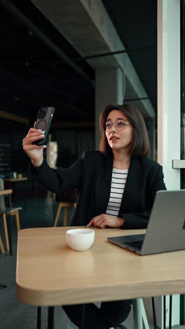 brunette woman wearing glasses, stripy shirt and black jacket speaks via video chat on her phone. business lady pointing and glancing at her laptop on the desk. vertical video.