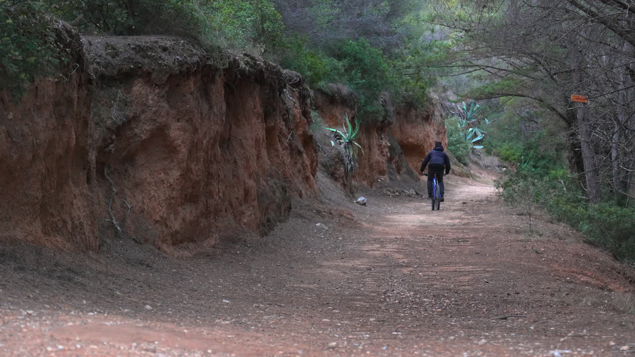 Mountain biker rides down dirt track along cliff wall and dense forest