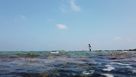 A-fisherman-stands-on-the-rocks-on-a-sunny-hot-day-at-high-tide-and-casts-a-hook-and-line-with-bait-again-and-again-to-catch-fish-to-eat-on-the-andaman-islands