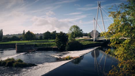 water cascades over the weir at miller's bridge on the river exe in exeter, devon with an autumn tree blowing in the foreground under a cloudy blue sky