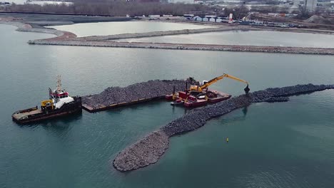 excavator on barge building breakwater from rocks near waterfront port