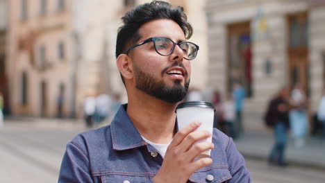 young indian man enjoying drinking morning coffee hot drink, relaxing, taking a break in city street