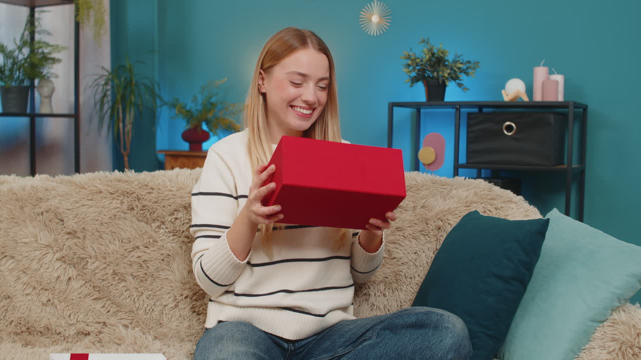 Caucasian woman opening birthday surprise gift on sofa smiling excited indoors happy joyful reaction
