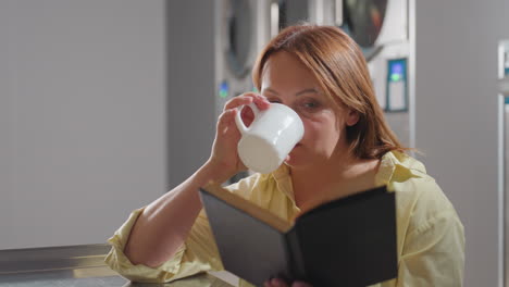 business woman seated over table inside laundromat reading notebook while sipping coffee from mock cup placed on surface, stainless washers in background glowing with digital panels, moment of work