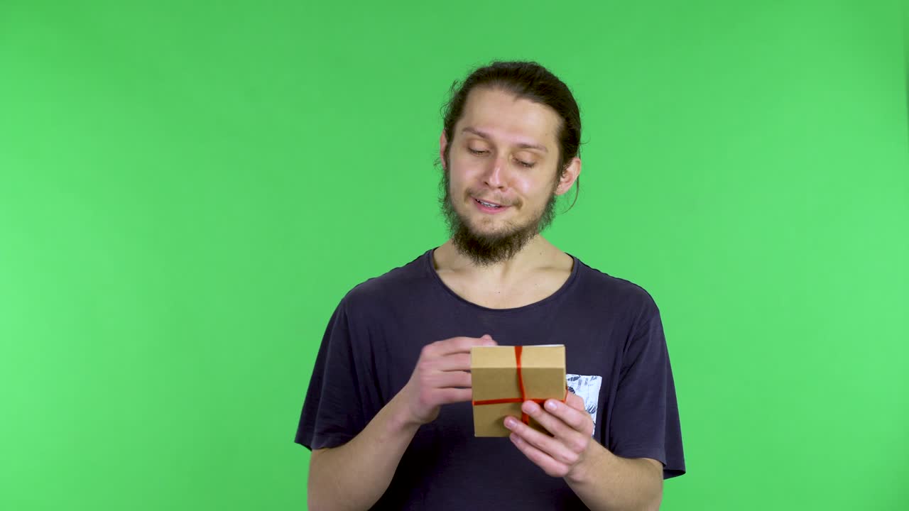 어두운 머리카락의 남자가 천천히 선물 상자를 열고 생일 놀라움을 펼치고 선물에 기하는 행복의 놀라움을 표현합니다.