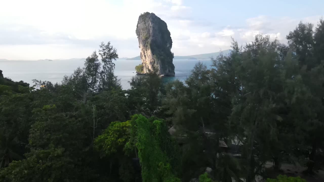 Beautiful tall palm trees with solar-powered houses between the trees near the beautiful lone high rock in the very clear blue sea at the touristic Ko Poda Beach. Drone tilt shot