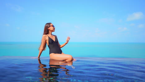 Sexy-asian-female-in-black-swimsuit-sitting-on-infinity-swimming-pool-border-and-enjoying-on-sunny-summer-day-and-her-tropical-holiday,-full-frame