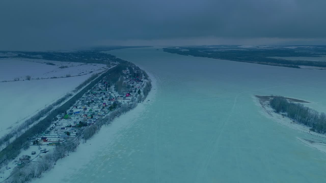 Hava görünümü, helikopter donmuş bir nehrin üzerinden uçuyor, boyunca bir köy uzanıyor. Buzla kaplı geniş nehir, kışın su alanları. Ayazlı kış günü. 4K, ProRes