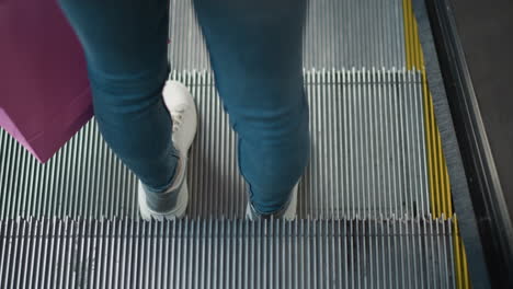 back view close-up of lady holding shopping bag standing on moving escalator in retail store, blue jeans, white sneakers, and pink bag create modern shopping vibe