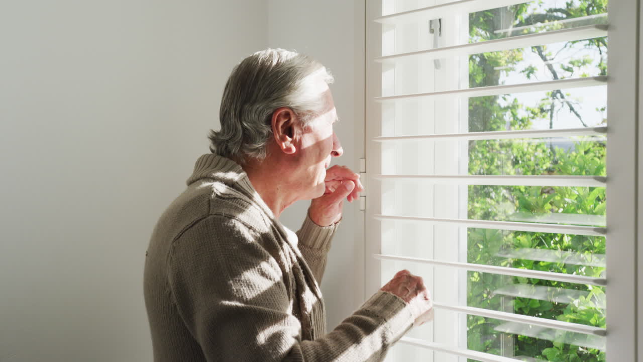 Retired senior man in cardigan sweater looking out through window blinds at home