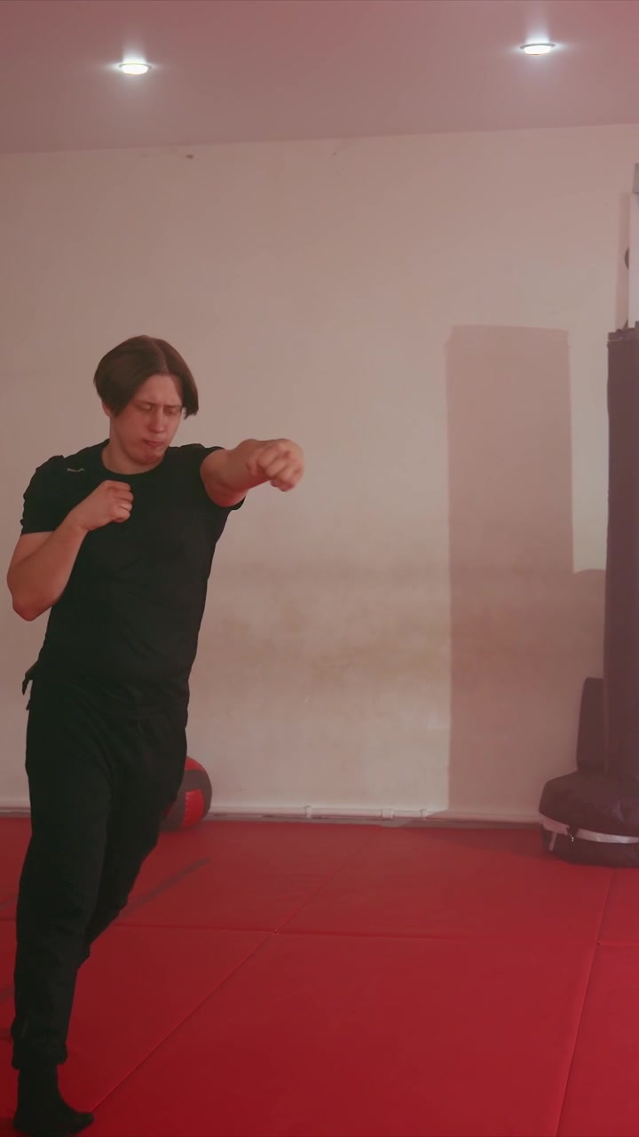 Young men in black clothing perform martial arts movements. They maintain fighting stances with fists raised interacting. White walls ceiling lights radiators medicine balls