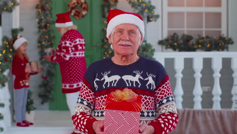 Portrait-of-grandfather-man-presenting-gift-box-smiling-near-decorated-Christmas-house-with-family