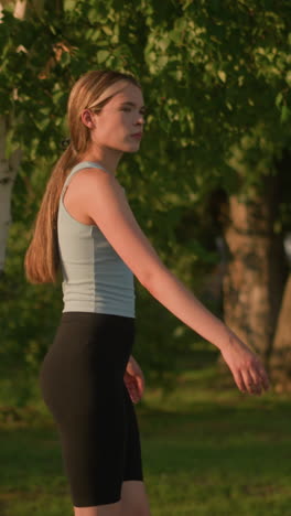 vista lateral de una joven que parece infeliz mientras se mueve con gracia como patinaje sobre ruedas al aire libre, la luz del sol se refleja en ella, el cabello atado hacia atrás, fondo natural con árboles verdes exuberantes