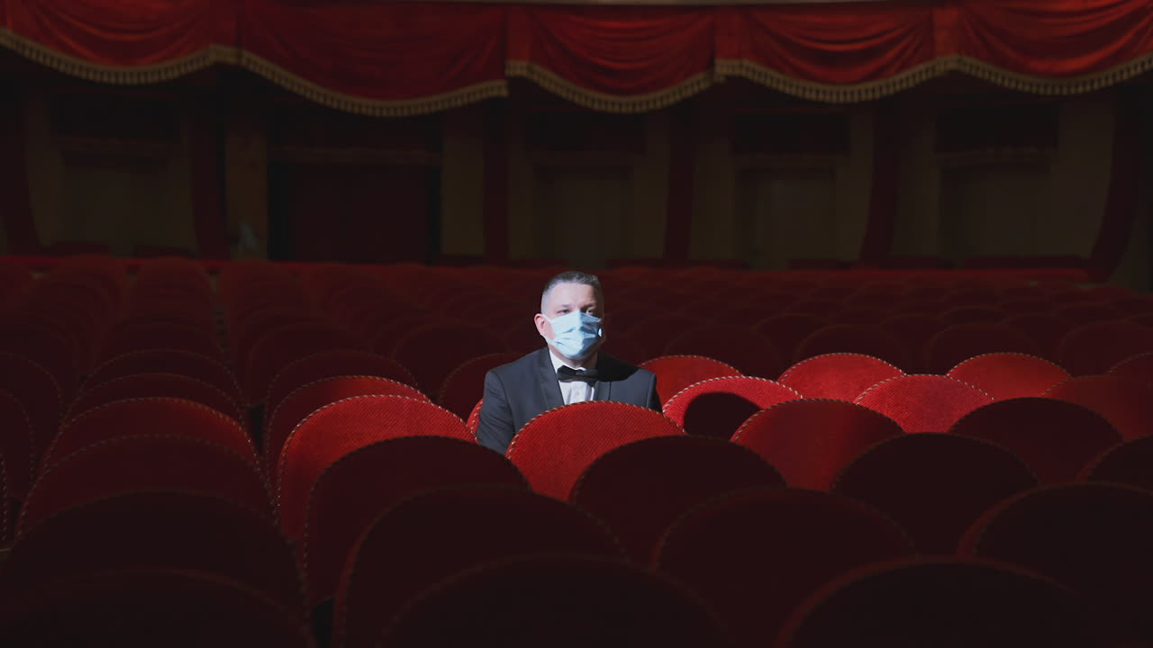 Lonely man sitting on theater seat. Adult man in medical mask watches in empty theater hall