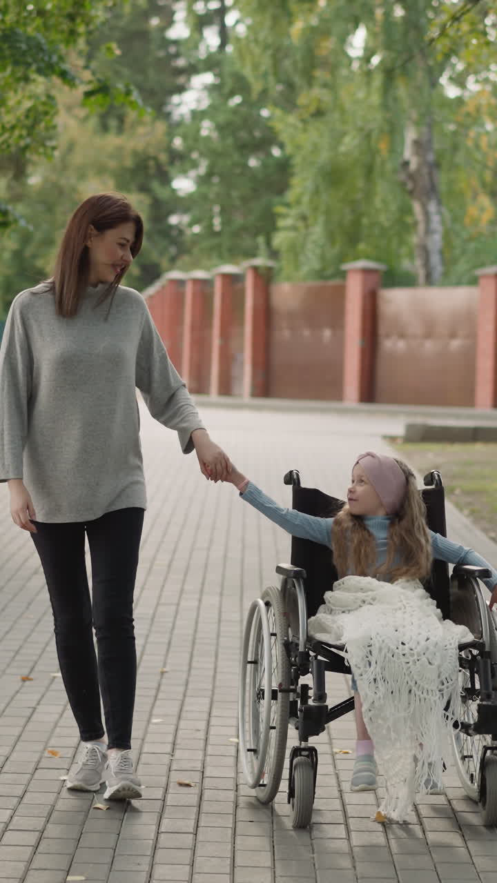 madre sostiene la mano de su hija en silla de ruedas caminando por la carretera en la parte de la ciudad. mujer joven y niña con lesión de la médula espinal disfrutan del fin de semana juntos