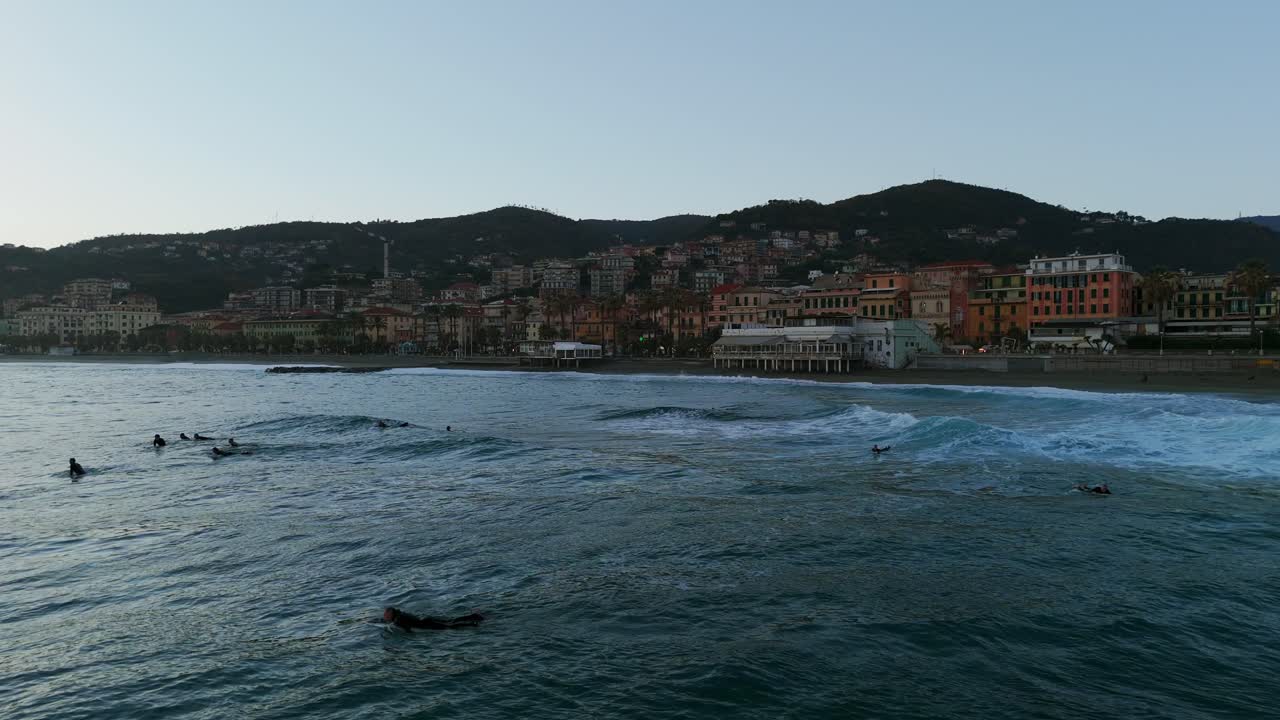 Gruppo di surfisti che cavalcano le onde al tramonto con la costa di Varazze sullo sfondo.