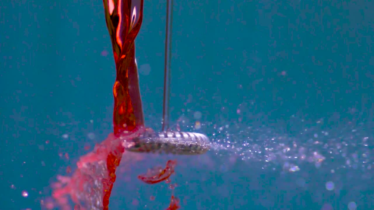 A small hand mixer or a blender being put near a stream of red water or fluid and then against it making the water stream splatter in slow motion. Footage is filmed on a blurry blue background.