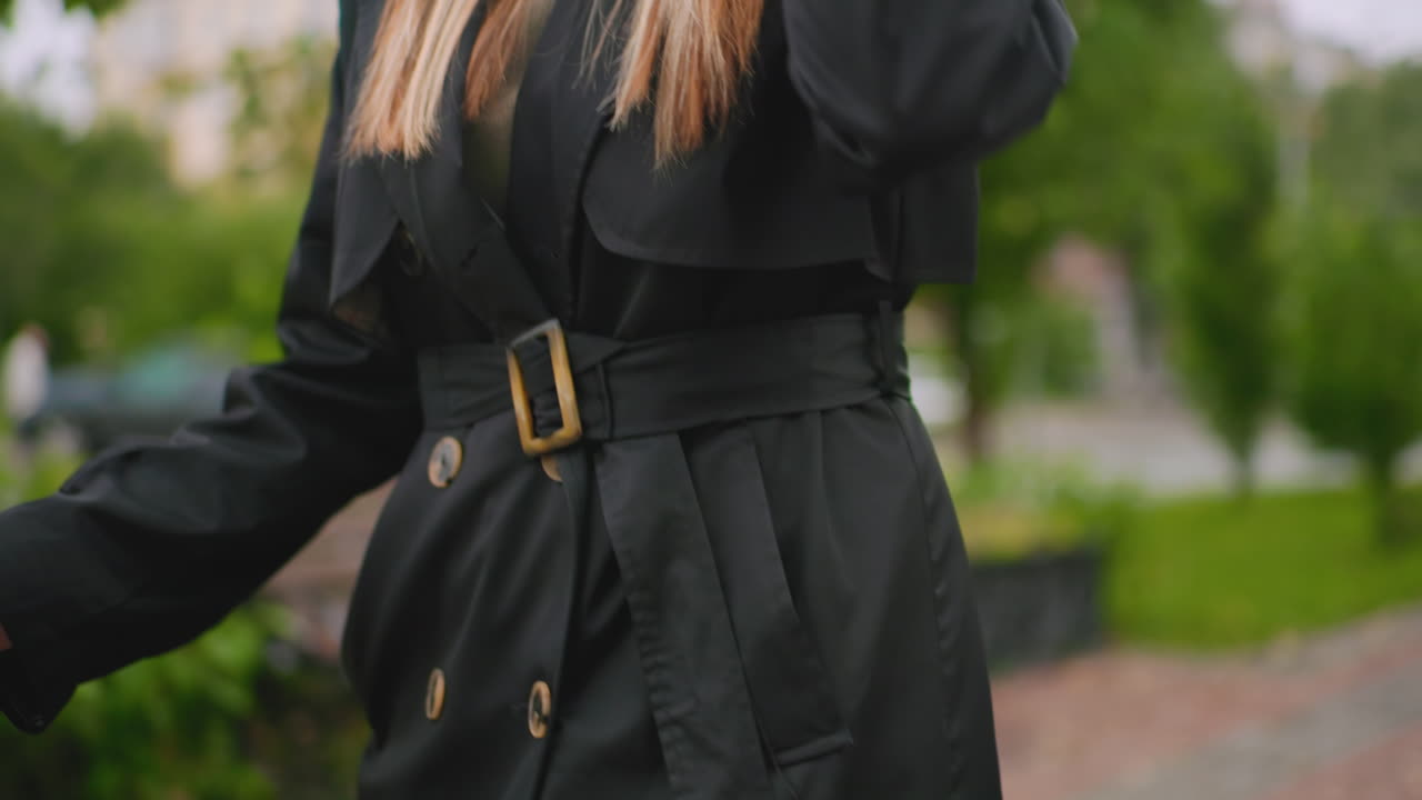 Close up of woman in black trench coat holding umbrella with wooden handle, detail of belt buckle and buttons visible, showing autumn fashion style during overcast day in outdoor urban park environment