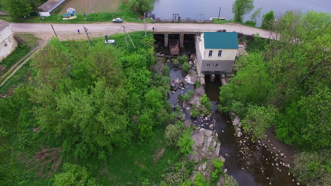 Quick flight over the narrow river with green banks, dam over it. Beautiful farmlands at backdrop. Aerial perspective.