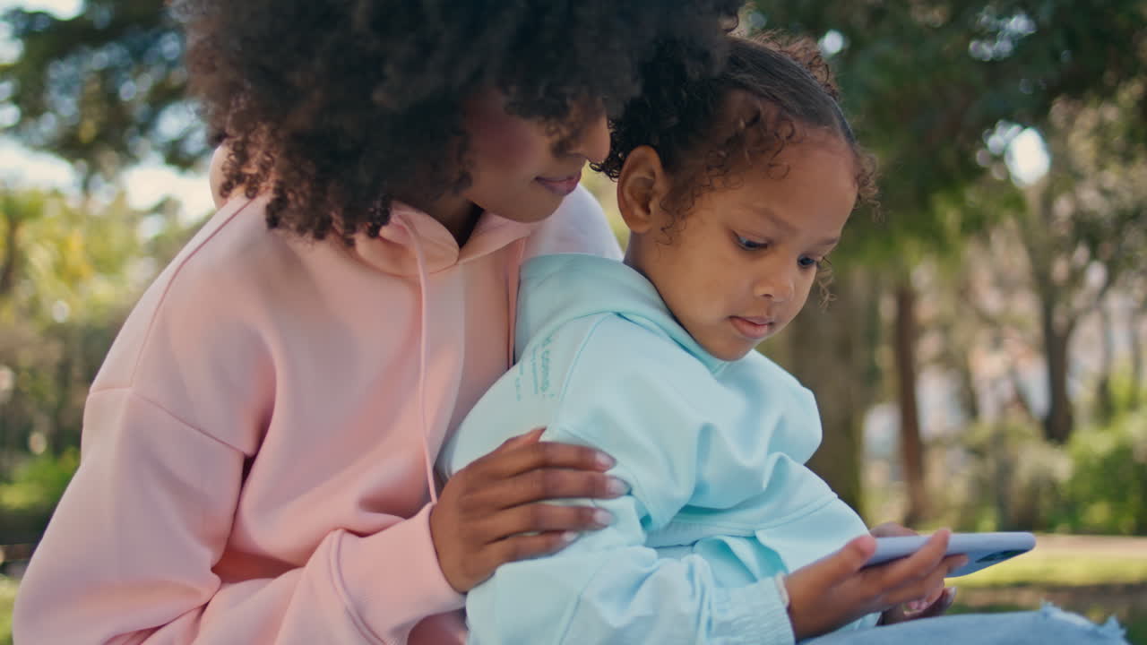 Little girl watching mobile phone sitting on green nature with mother close up.
