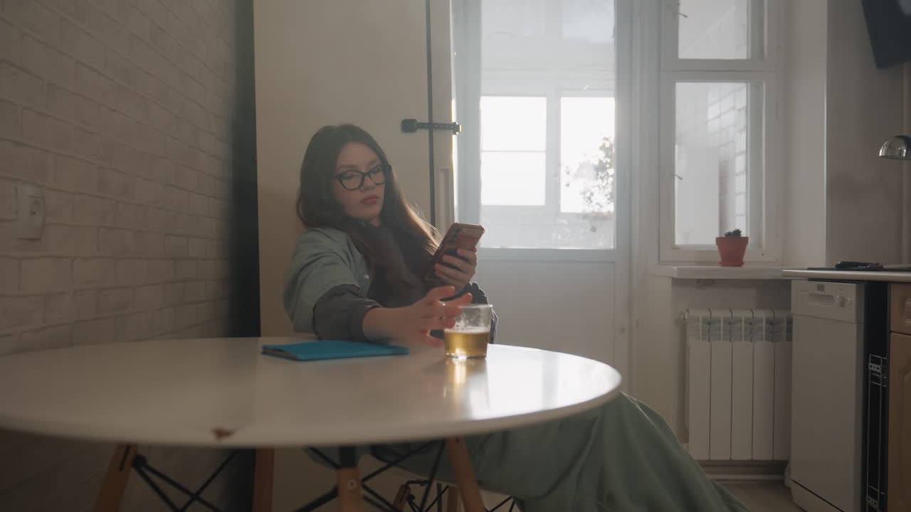 Giovane donna bianca al tavolo della cucina, appoggiata all'indietro, scorrendo il telefono e sorseggiando tè, morbida luce mattutina dalla finestra, interno accogliente di un appartamento, studentessa freelance che si prende una pausa tranquilla, umore domestico pensieroso
