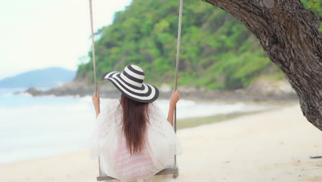 back of young woman on a tree swing by exotic tropical white sand beach and sea, luxury vacation concept, slow motion full frame