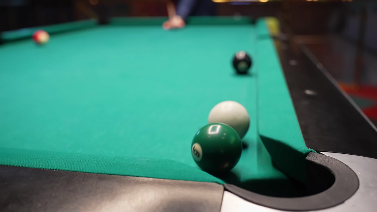 Close-up of a green billiard ball rolling into a corner pocket on a pool table, with cue ball and other balls on green felt, filmed indoors with shallow depth of field.