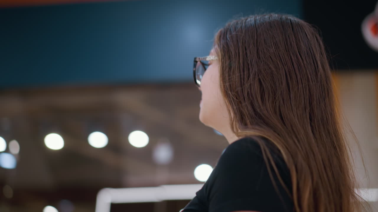 Close-up side view of woman wearing glasses in black top looking up with soft bokeh lights in background, blurred inscription on wall adds depth to modern urban retail environment
