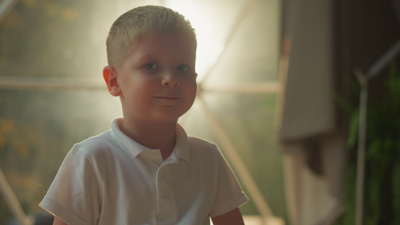 조명 된 글램핑 텐트 근처의 유치원 소년의 초상화. 즐거운 아이는 관광 여행을 즐기고 웃으며 자연 장소를 근접합니다. 작은 하이커는 모호한 배경에서 저녁에 휴식을 취합니다.