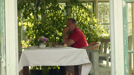 man sits at table in a sunlit greenhouse surrounded by plants, enjoying a quiet moment