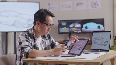 vista lateral de un hombre asiático comparando el diseño de su coche en una tableta con la foto en su teléfono inteligente mientras trabaja en el estudio con tv y computadoras muestra modelo de coche eléctrico 3d