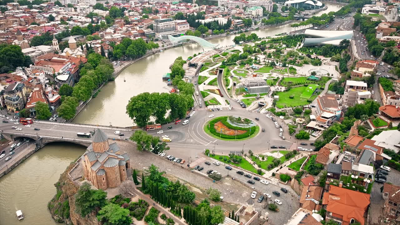 Aerial drone view of Tbilisi, Georgia at cloudy weather. Metekhi Church, Rike Park, water channel, multiple buildings, a lot of greenery, road with traffic