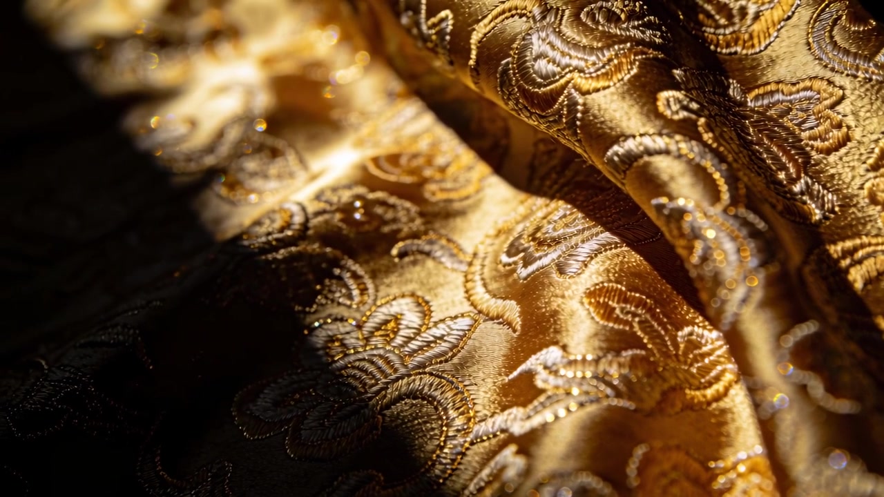 Un raggio caldo si muove attraverso un primo piano di tessuto broccato dorato, creando riflessi metallici.
