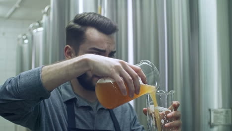 Joven-Y-Alegre-Maestro-Cervecero-Mirando-Un-Vaso-De-Cerveza-Sin-Filtrar-Recién-Hecha-Y-Vertiéndola-En-Otro-Recipiente