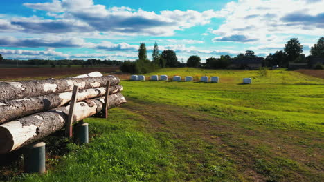 Dry-logs-stacked-in-a-pile-on-ramp-next-to-a-sawmill,-countryside-scene-with-fodder-rolls,-aerial-dolly-out