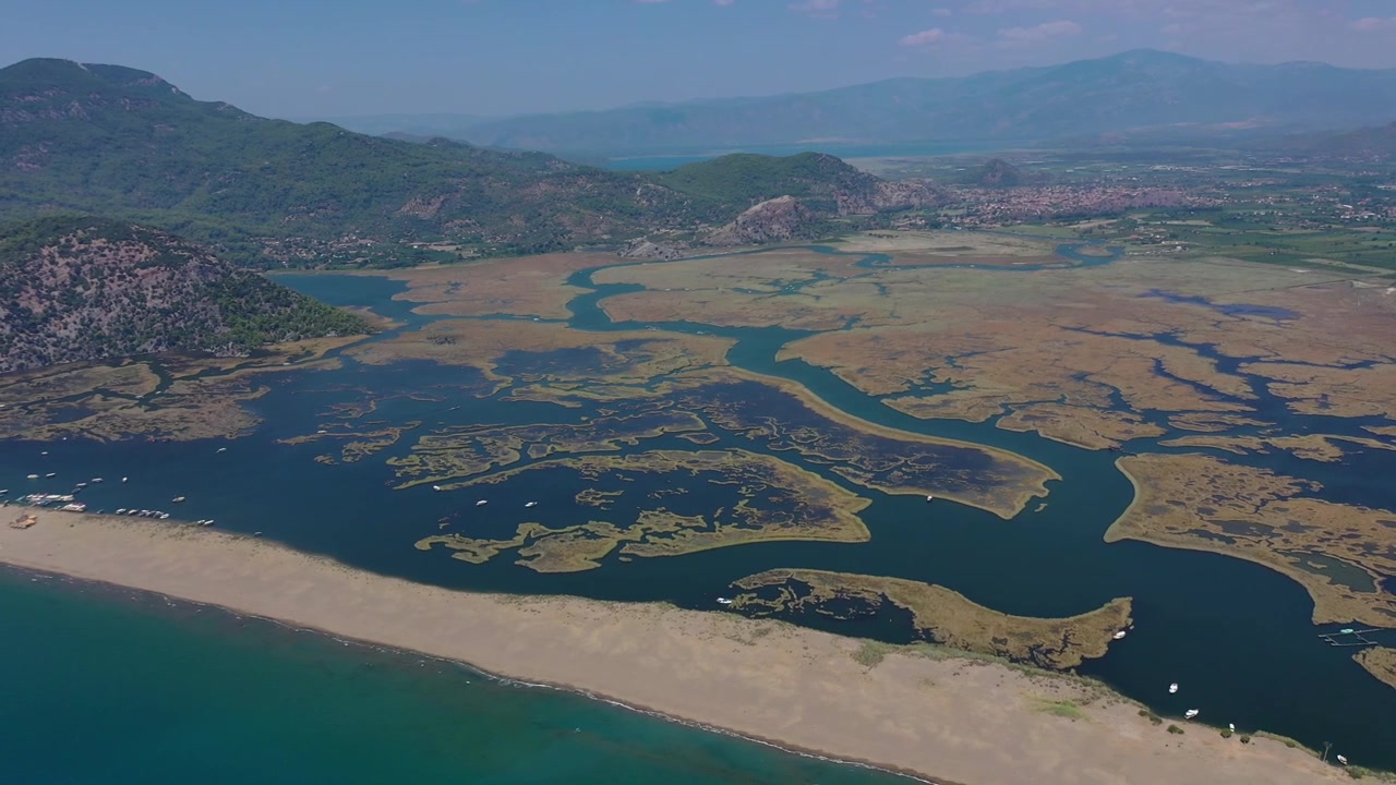 Muğla, Türkiye'de, Dalyan Nehri sazlık deltasının turkuaz Akdeniz ile buluştuğu İztuzu Plajı'nın yeşil dağlarla çevrili, açık ve güneşli bir günde yüksekten çekilmiş havadan drone panoraması.
