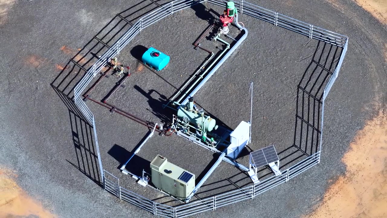 High-angle drone view rotating above a fenced industrial gas well facility in bright daylight.