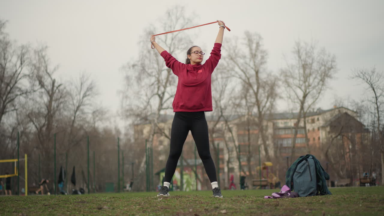 White Woman Swinging Triangular Loop With Wide Arcs Performing Dynamic Training Moves On Grass, Energetic Motion, Red Hoodie, Urban Playground In Soft Cloudy Light, Athletic Focus
