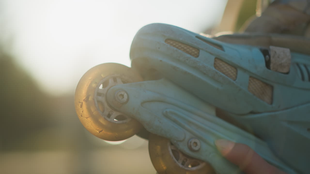 A close-up shot of a light blue rollerblade being held by a hand in a sunlit park. The image focuses on the rollerblade's wheels, capturing the details under warm sunlight