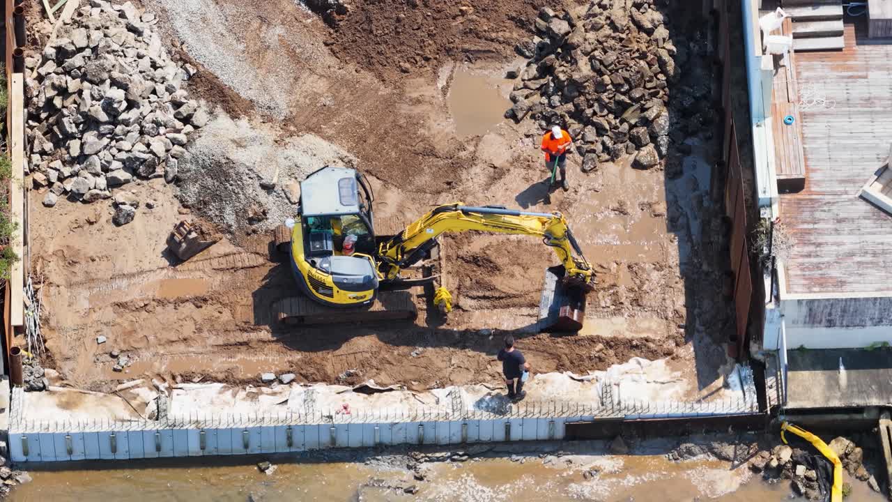 Drone overhead shot of mini excavator and workers digging muddy waterfront foundation in bright daylight