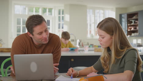 Padres-Ayudando-A-Su-Hija-Adolescente-Con-La-Tarea-Sentados-En-La-Mesa-De-La-Cocina-En-Casa-Usando-Una-Computadora-Portátil---Filmados-En-Cámara-Lenta