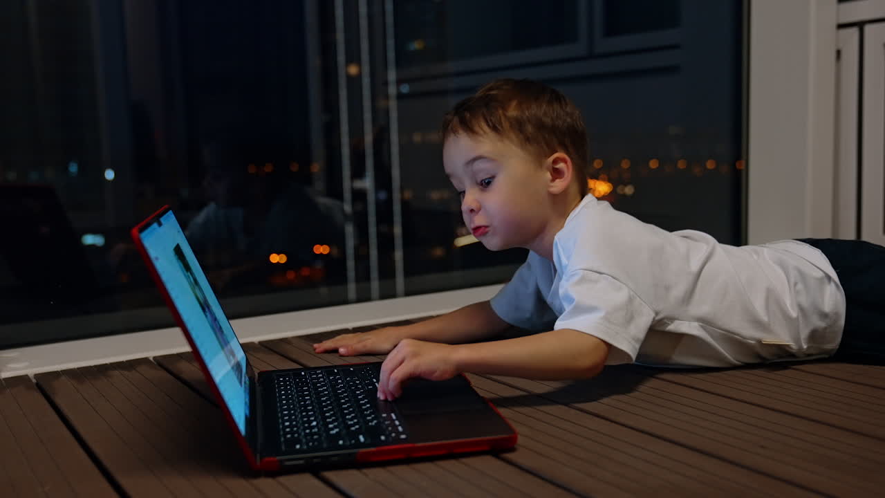 Child lying on the floor with laptop at night. A boy lies on the floor at night in front of a glowing laptop, with the city lights reflecting through the window