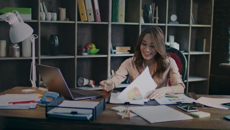 Smiling-businesswoman-reading-documents-with-good-statistics-graph-at-desk