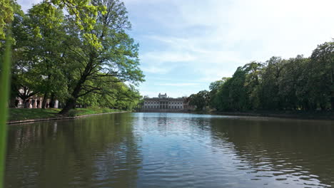 view of pałac na wyspie in warsaw with a serene lake and green trees in the foreground