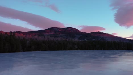 aerial slide flying low across the surface of a frozen pond looking at a mountain lit by sunrise