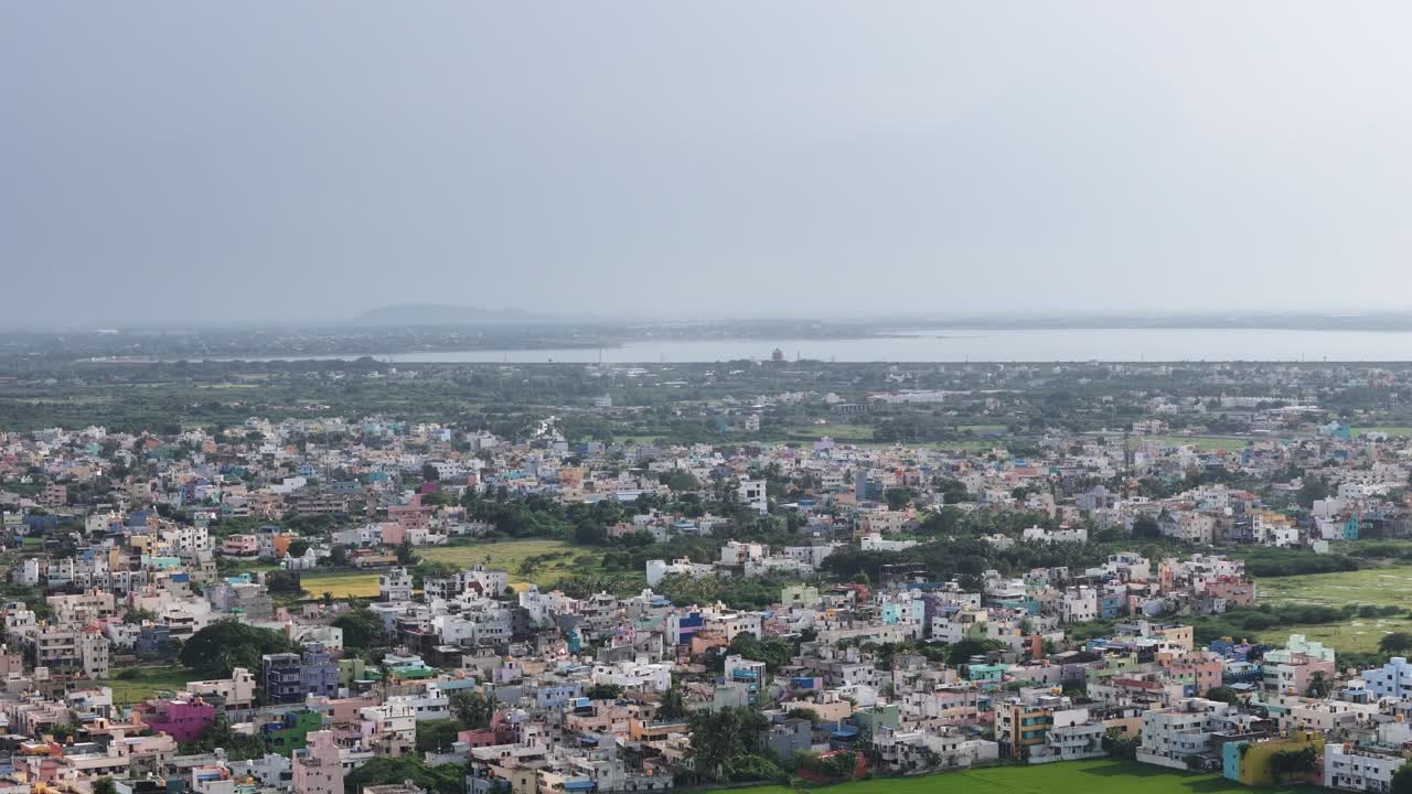Una vista aerea sfocata e grandangolare di edifici residenziali colorati, bassi e medi, caratteristici della periferia di Chennai. Gli edifici sono molto vicini tra loro e si estendono verso un lago distante o un grande specchio d'acqua.