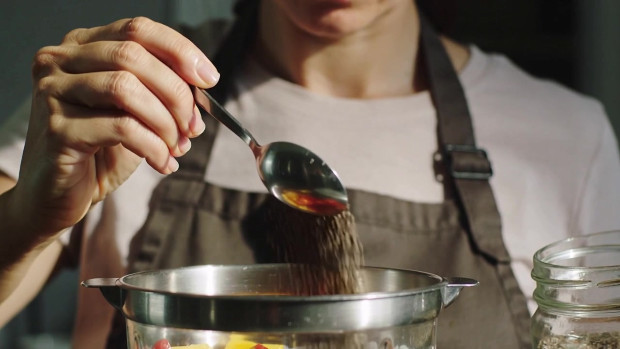 Holding jar, woman in dark apron scooping seeds with spoon, sprinkling to top yogurt and fruit