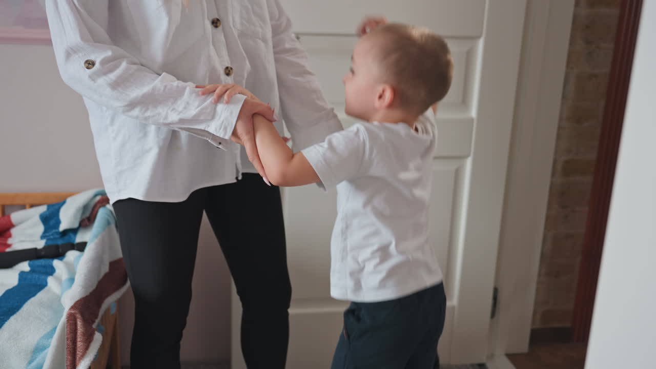 White mom and son tugging hands in bedroom doorway, playful boy reaching up while mother gently holds wrist, casual white shirt, colorful quilt on bed, warm natural light, everyday family bonding